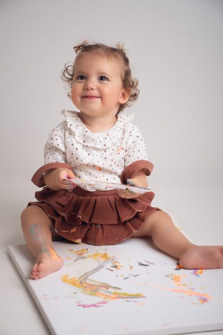 Ein lächelndes Kleinkind sitzt auf dem Boden und spielt mit Farben auf Papier.