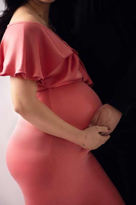 Schwangere Frau in figurbetontem, rosafarbenem Kleid umarmt ihren Bauch.
