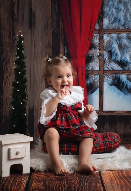 Mädchen in kariertem Kleid sitzt barfuß vor einem Fenster mit Schnee und einem kleinen Baum.