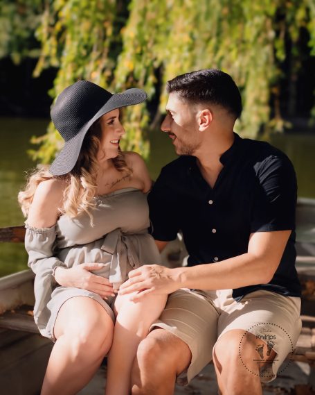Paar sitzt am Wasser, lächelt sich an, die Frau trägt einen großen Hut.