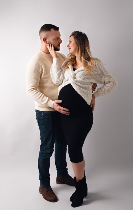 Schwangere Frau und Mann posieren liebevoll auf neutralem Hintergrund.