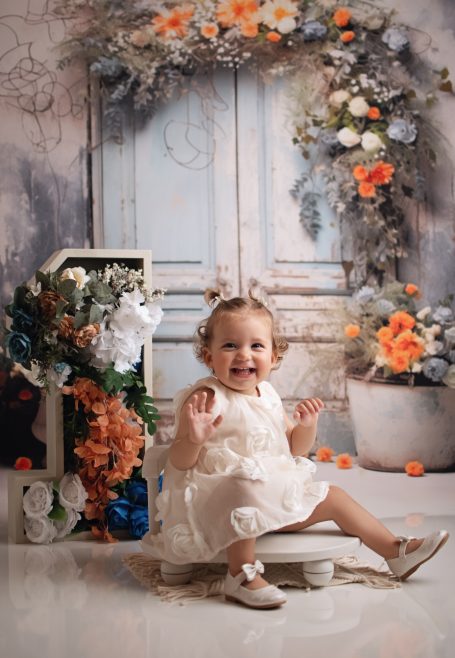 Ein lächelndes kleines Mädchen in einem weißen Kleid sitzt vor einer blumengeschmückten Kulisse.