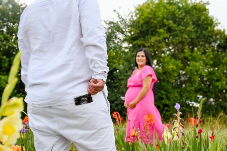Schwangere Frau in pinkem Kleid steht in Blumenfeld, Mann blickt zu ihr.