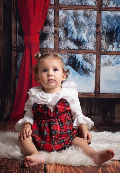 Ein kleines Kind in einem roten karierten Kleid sitzt auf einem flauschigen Teppich.