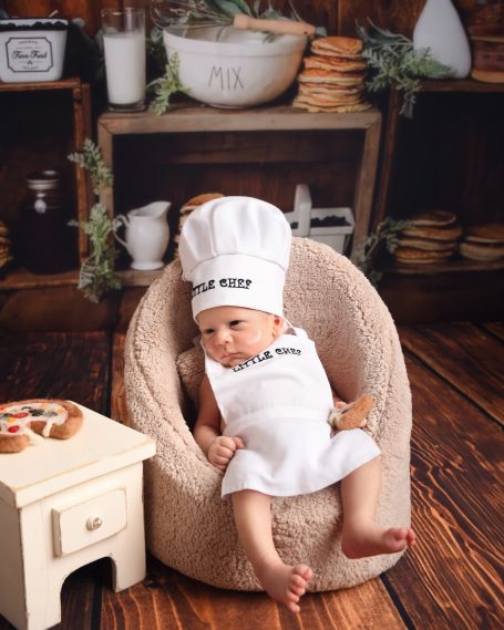 Ein Baby im Kochoutfit sitzt in einem beigen Sessel mit Küchenutensilien im Hintergrund.
