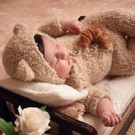 Süßes Baby in einem braunen Bärenanzug, schlafend auf einem Bett mit Spielzeug.