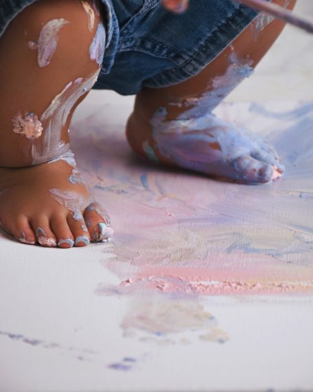 Barfußkind mit bemalten Füßen und Händen, das auf einem Malboden steht.