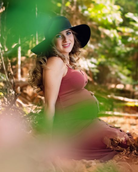 Schwangere Frau in einem roten Kleid und schwarzem Hut, umgeben von Natur.