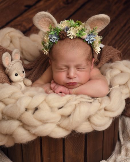 Ein schlafendes Baby mit Hasenohren und Blumenkranz, umgeben von weichem Material.