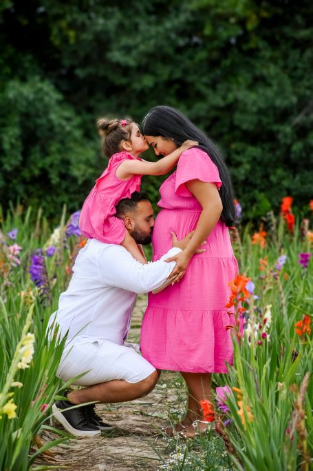 Familienfoto: Mutter in pinkem Kleid, Kind auf den Schultern, Vater kniet im Blumenfeld.