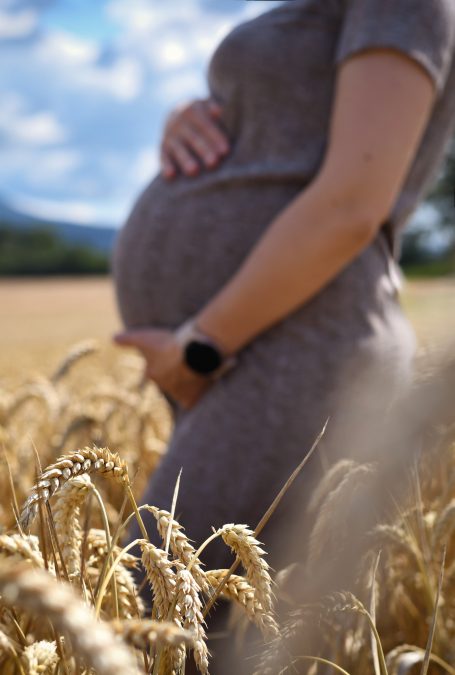 Schwangere Person in einem Weizenfeld mit blauem Himmel im Hintergrund.