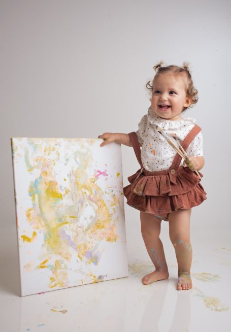 Ein lächelndes Kind steht neben einer bemalten Leinwand und hält einen Pinsel.