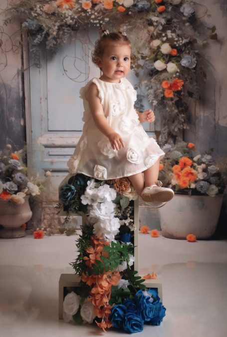 Kleines Mädchen in weißem Kleid sitzt auf einem blumenverzierten Podest.