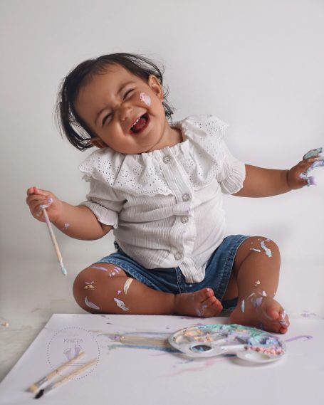 Ein lachendes Kleinkind mit Farbstiften, schmutzig von Farbe, sitzt auf dem Boden.