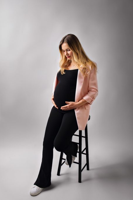 Schwangere Frau in schwarzem Outfit sitzt auf einem Hocker und hält ihren Bauch.