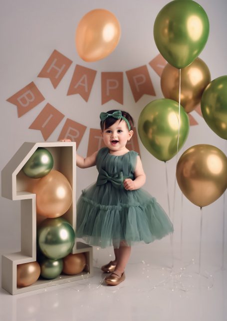 Ein kleines Mädchen in einem grünen Kleid steht neben einem „1“-Schild und Luftballons.