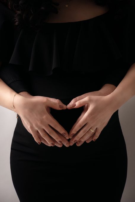 Hände formen ein Herz über einem schwangeren Bauch in eleganter schwarzer Kleidung.