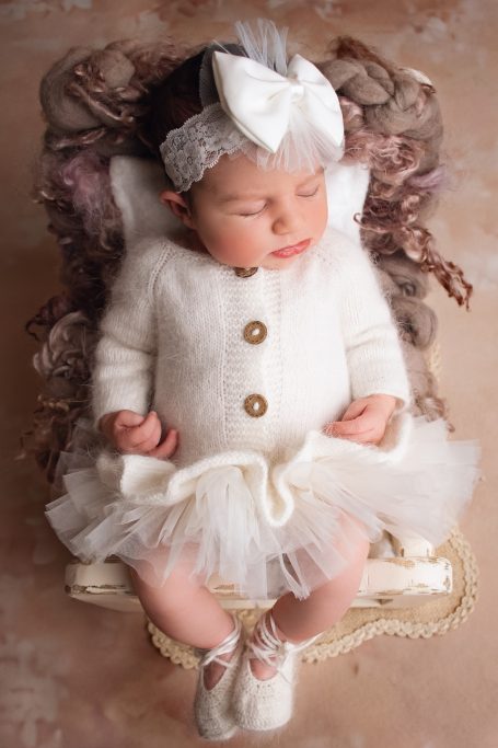 Neugeborenes Mädchen in weißem Kleid mit einer großen Schleife auf dem Kopf, schlafend.