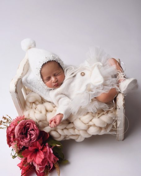 Ein liegendes Baby in einem weißen Strampler, umgeben von rosa Blumen.