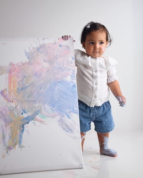 Ein Kleinkind steht neben einer bemalten Leinwand und hat Farben an Händen und Füßen.