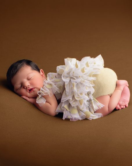 Ein schlafendes Baby in einem weißen, fransigen Outfit auf einem braunen Hintergrund.