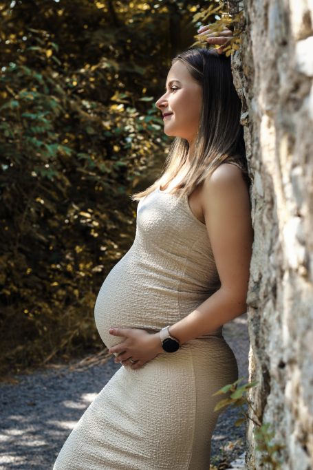 Schwangere Frau in einem engen Kleid, die an einem Baum steht und ihren Bauch hält.