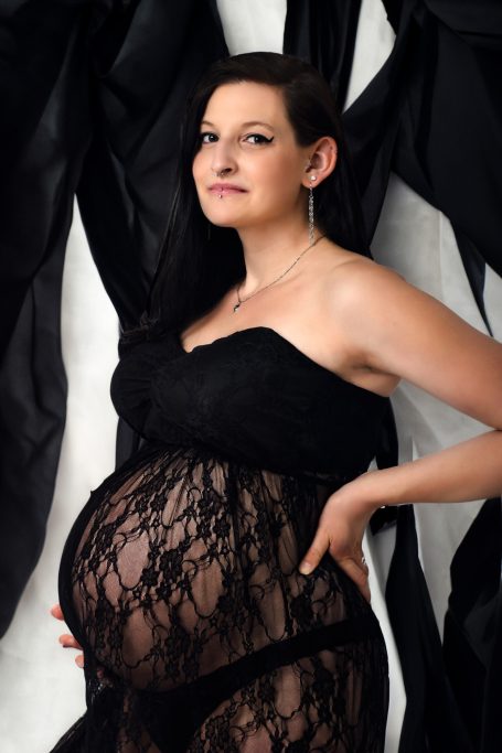 Schwangere Frau in schwarzem Spitzenkleid vor schwarzem Hintergrund.
