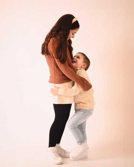 Junge umarmt lachend eine stehende Frau in heller Kleidung vor neutralem Hintergrund.