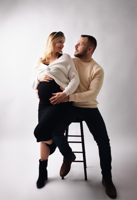 Schwangere Frau sitzt auf einem Stuhl, umarmt von einem Mann in casual Kleidung.