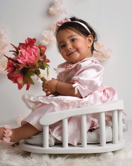 Kleines Mädchen in rosa Kleid sitzt auf einem Schaukelstuhl und hält einen Blumenstrauß.