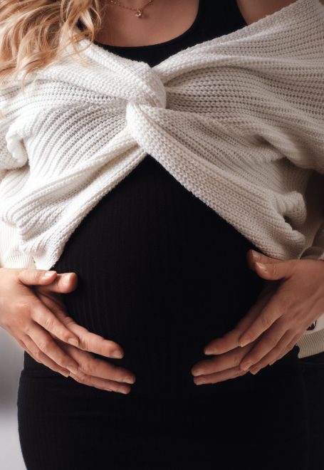 Schwangere Person mit ihren Händen auf dem Bauch, in einem schwarzen Outfit und heller Jacke.