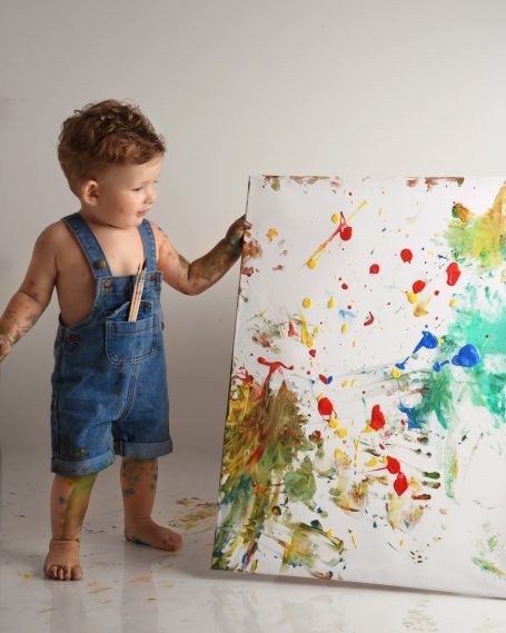 Kleines Kind in Latzhose hält bemalte Leinwand mit bunten Farbklecksen.