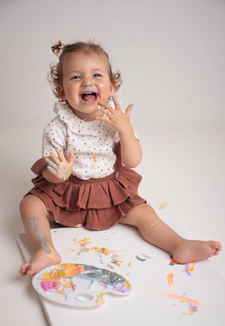 Ein lächelndes Kleinkind mit Fingerfarbe sitzt auf dem Boden und spielt mit buntem Papier.