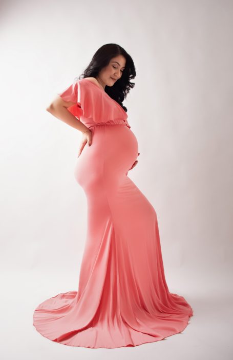 Schwangere Frau in einem eleganten, rosa Kleid, die lächelnd ihre Hand auf den Bauch legt.